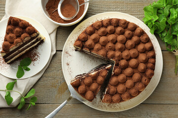 Delicious tiramisu cake, cocoa powder, server and mint leaves on wooden table, flat lay