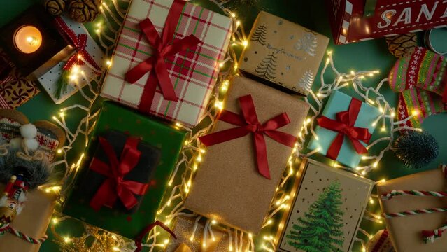 Top view on green background decorated with gift boxes, Christmas decorations and glowing garlands. Golden Christmas lights illuminates present tied with red satin ribbons, rotating shot on RED camera