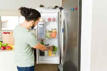 Latin young man opening the fridge in the kitchen and organizing the groceries while doing house...