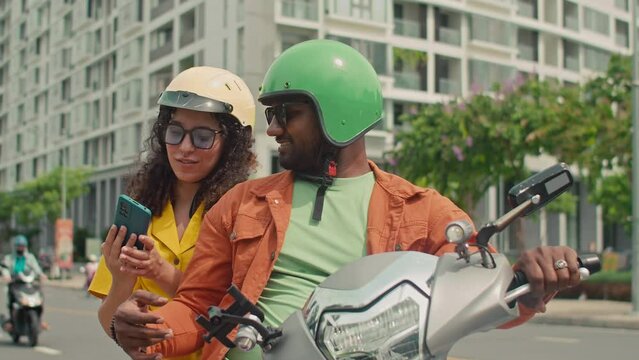 Medium shot of African American husband and biracial wife in helmets sitting on scooter in city street, taking selfies on smartphone in front of skyscraper, posing and smiling, then looking at photos