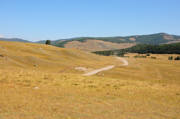 A winding field road going along the gentle slope of the hills in the autumn steppe to meet a high mountain range.