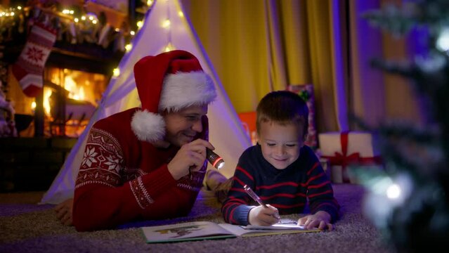 Cute little funny kid son making wishes and writing letter to Santa Claus. Father with child boy bonding fun playing together 4K. Happy family loving dad in Santa red hat with flashlight with kid