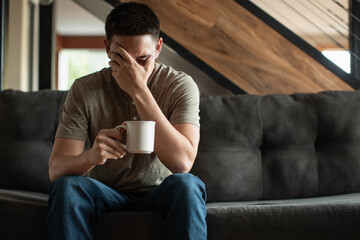 exhausted, tired, and stressed man sitting on sofa drinking coffee - obrazy, fototapety, plakaty