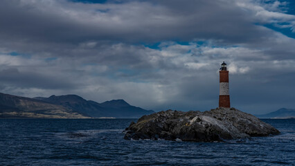 The famous southernmost old lighthouse Les Eclaireurs  in the Beagle Channel. A stone tower with red and white stripes stands on a rocky island against the sky and clouds. Mountains in the distance