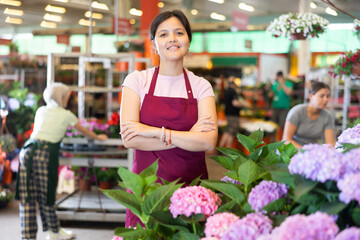 Smiling middle-aged saleswoman standing next to the counter of hydrangea flowers in open-air plants...