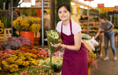 Smiling polite young asian female florist in apron working at flower market, offering bouquet of delicate blooming daisies..