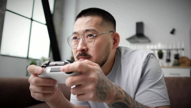 Asian Man Playing Video Game Holding Joystick Close Up. 30s man playing games with controller sitting on sofa at home. Handheld Camera. 