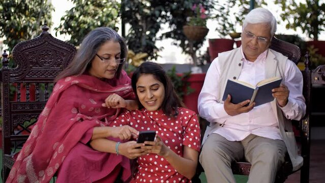 An elderly woman and her daughter use a mobile while her husband is reading a book - family bonding  family union. A beautiful girl in casual clothes is spending time with her parents - Indian fami...