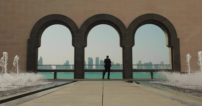 Business trip to Doha. A man in suit taking photos of Doha skyline from Museum of Islamic Art courtyard with fountains, Doha, Qatar - June 20, 2023