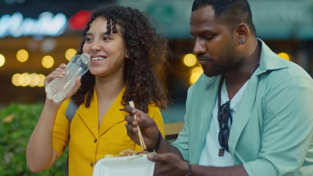 Medium close-up shot of African American husband and biracial wife eating delicious exotic street food while travelling abroad, exchanging impressions, unimpressed girl shaking head and drinking water