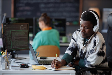 Serious programmer in headphones making notes in notebook while working with security codes on...