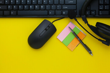 A Black keyboard, wired mouse, black headphones, sticky notes, and a pen on a Yellow Background....
