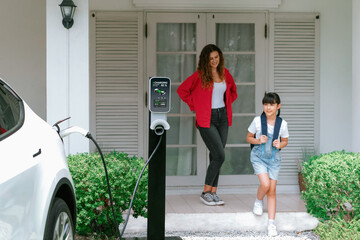 Happy little young girl learn about eco-friendly and energy sustainability as she help her mother recharge electric vehicle from home EV charging station. EV car and modern family concept. Synchronos - obrazy, fototapety, plakaty