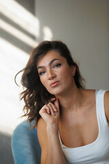 Beautiful brunette woman in white top and blue jeans sitting. Girl smile, happy. Portrait of young pretty woman. Smile and freshness. White background. Light from window, shadows