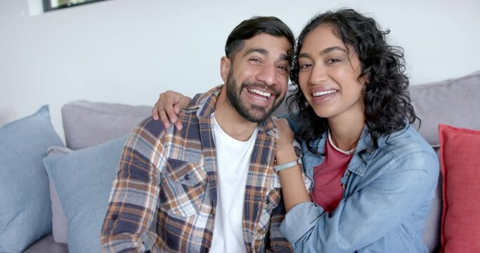 Portrait of happy biracial couple sitting on sofa embracing and smiling at home, in slow motion