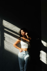 Beautiful brunette woman in white top and blue jeans. Girl smile, happy. Portrait of young pretty woman. Complicated sunlight with shadows. White background. Light from window, portrait.