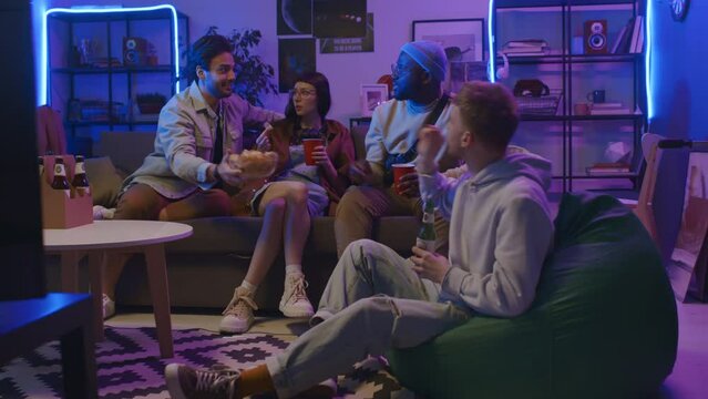 Full shot of four diverse casually dressed young people relaxing on couch in living room with blue and pink neon lighting, drinking beer, sharing potato crisps and chatting