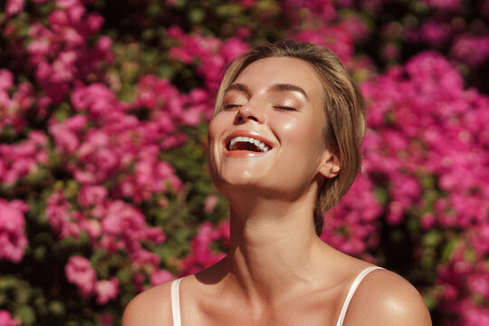 Beautiful blonde woman with smooth skin and natural makeup, as she poses against a backdrop of a bush with delicate pink flowers