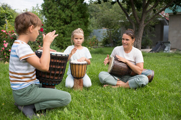 mom and two kids play at ethnic drums sitting on the grass on a sunny day. a family hobby. children...