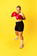 Full length portrait of young smiling chubby sports woman in tracksuit and red boxing gloves isolated over yellow background.