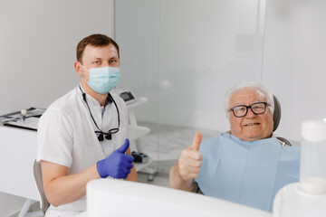 In front of the camera at the dentist office the doctor and patient posing in front of the camera charismatic old man patient laying down on the dentist chair the doctor wearing the protective mask - Powered by Adobe