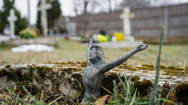 old damaged metal figurine of Christ with pierced hands, placed on a tombstone, looks like making a gesture of victory and a cross in the background