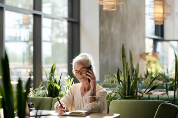 Senior woman having conversation on mobile phone and making notes in her notepad during her work in...