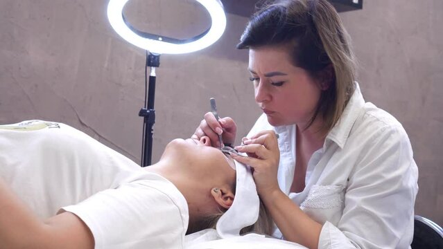 Video of process of eyelash extension, young lashmaker sitting over client lying on couch and gluing fake lashes on lid using tweezers. Concentrated master holding tools and touching cilia.