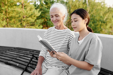 Nurse reading book to senior woman while they sitting on bench outdoors