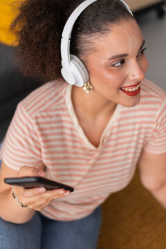 High angle of positive young female in casual clothes listening to music in wireless headphones and browsing mobile phone while looking at away