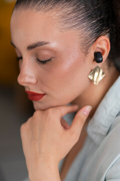 Crop side view of young thoughtful female with makeup touching chin while resting and listening to music over wireless ear bud in room against blurred background
