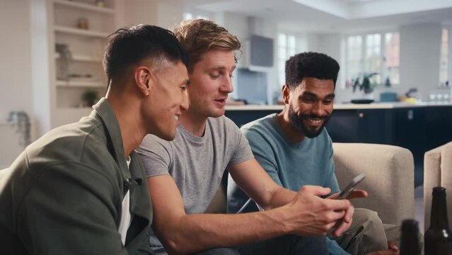 Group of male friends sitting on sofa at home looking at dating app on mobile phone together - shot in slow motion