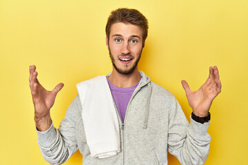 Sporty man in sweatshirt and towel on yellow receiving a pleasant surprise, excited and raising hands.