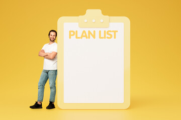 Positive confident millennial european man in casual with crossed arms on chest near big plan list with free space - obrazy, fototapety, plakaty