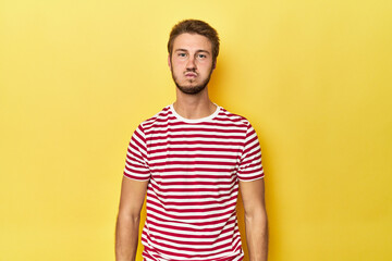 Young Caucasian man on a yellow studio background blows cheeks, has tired expression. Facial...