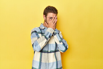 Young Caucasian man on a yellow studio background blink through fingers frightened and nervous.