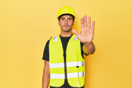 Latino worker in yellow vest and helmet standing with outstretched hand showing stop sign, preventing you.