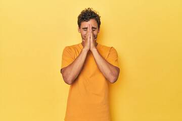 Young Latino man posing on yellow background holding hands in pray near mouth, feels confident.