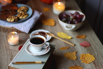 Cup of tea or coffee, plate with desserts, dried oranges, bowl of grapes, scented candles, vintage books, pumpkins and autumn leaves on the table. Autumnal hygge. Selective focus.