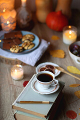 Cup of tea or coffee, plate with desserts, dried oranges, bowl of grapes, scented candles, vintage books, pumpkins and autumn leaves on the table. Autumnal hygge. Selective focus.