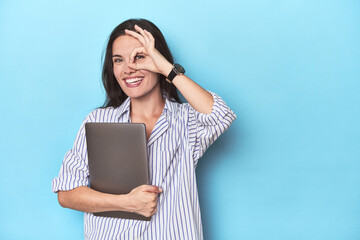 Young businesswoman holding laptop on blue excited keeping ok gesture on eye. - obrazy, fototapety, plakaty