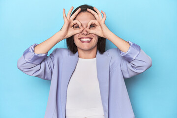 Woman in blue blazer on blue background showing okay sign over eyes
