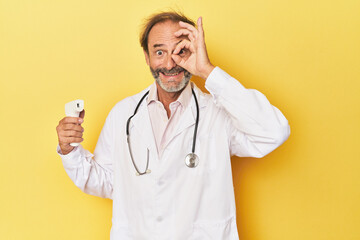 Doctor holding infrared thermometer in studio excited keeping ok gesture on eye.