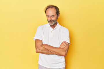 Caucasian middle-aged man in yellow studio frowning face in displeasure, keeps arms folded.