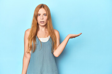 Fototapeta premium Redhead young woman on blue background impressed holding copy space on palm.