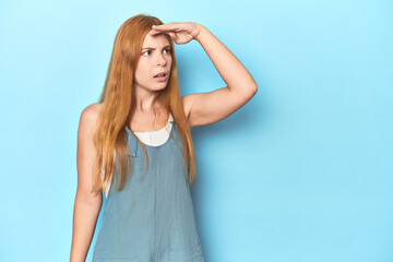 Redhead young woman on blue background looking far away keeping hand on forehead.