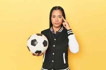 Indonesian schoolgirl with soccer ball on yellow pointing temple with finger, thinking, focused on a task.