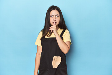 Young woman in kitchen apron on blue keeping a secret or asking for silence.