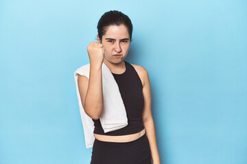 Fototapeta na wymiar Sporty woman with towel on blue studio showing fist to camera, aggressive facial expression.
