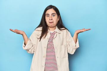 Young Caucasian woman on blue backdrop doubting and shrugging shoulders in questioning gesture.
